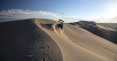 A vehicle climbs a sandy dune, kicking up dust against a backdrop of blue sky and distant hills.
