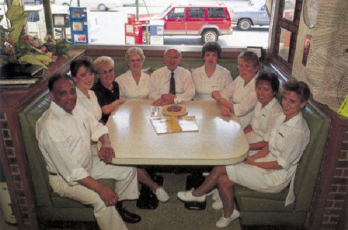 A group of nine people in white attire sitting around a table in a diner, smiling at the camera.
