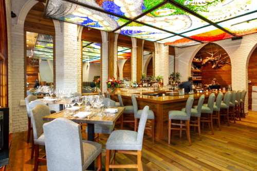 A stylish restaurant interior featuring a bar, wooden tables, gray chairs, and a colorful stained glass ceiling.
