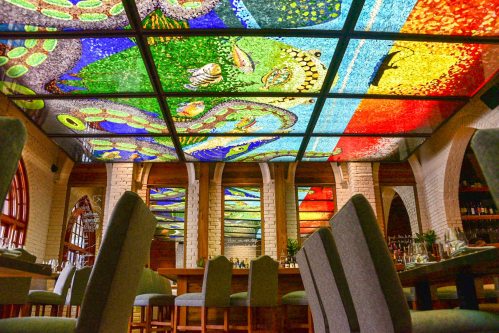 Colorful stained glass ceiling in a restaurant, with a view of tables and chairs below.