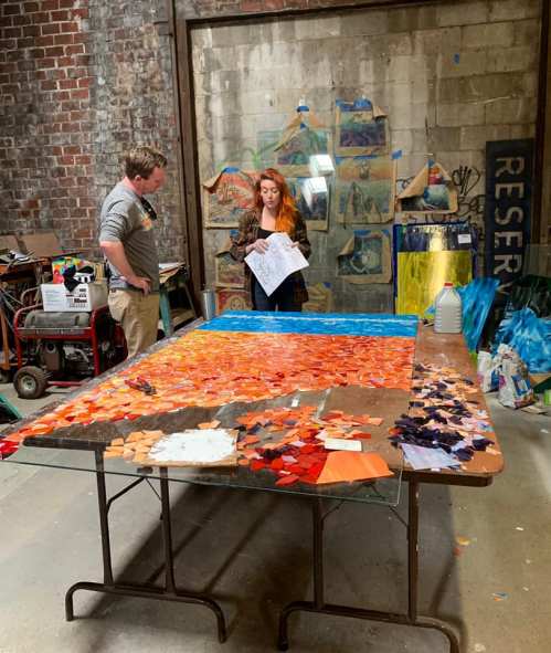 Two artists discuss a colorful mosaic project in a studio, surrounded by art supplies and a textured wall.