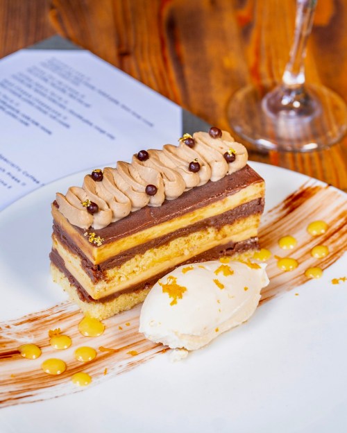 A layered chocolate cake with cream frosting, garnished with chocolate pearls, served with a scoop of vanilla ice cream.