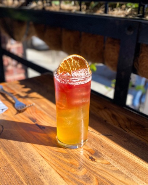A tall glass of layered iced tea with a slice of dried orange on top, sitting on a wooden table.