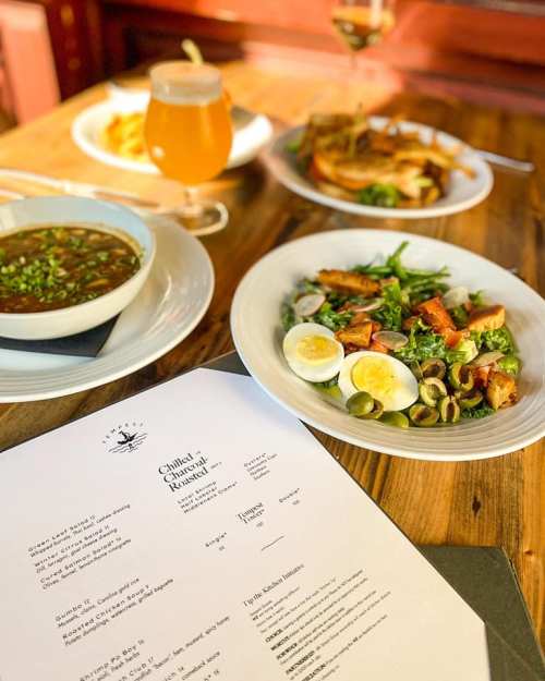 A table setting with a menu, a bowl of soup, a salad with eggs and chicken, and a glass of beer.