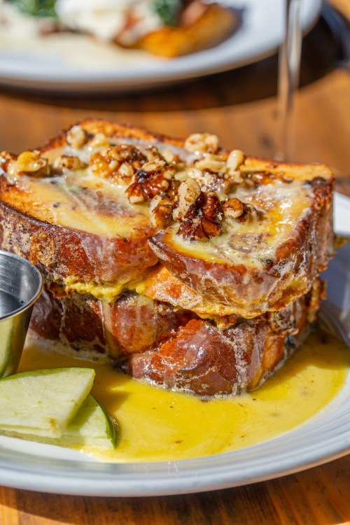 A close-up of French toast topped with nuts, served with a side of apple slices and syrup in a small cup.