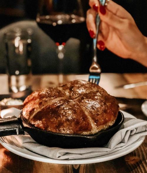 A hand with red nails holds a fork above a golden-brown bread in a cast-iron skillet on a table.