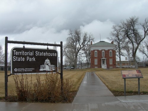 Sign for Territorial Statehouse State Park with a historic building in the background under a cloudy sky.