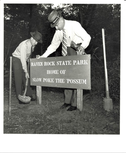 Two men install a sign for Raven Rock State Park, featuring "Home of Slow Poke the 'Possum," in a wooded area.