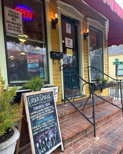 A colorful deli entrance with a neon "OPEN" sign, menu board, and potted plants on the steps.