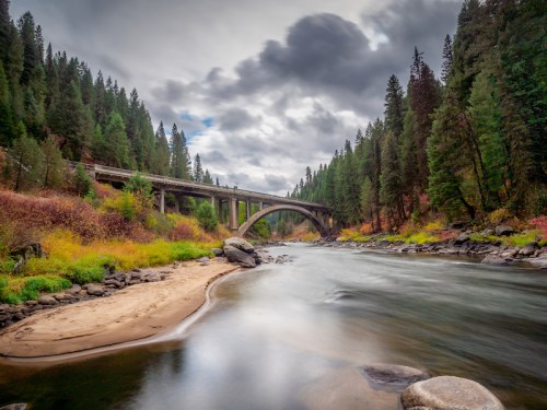 A serene river flows under a bridge, surrounded by lush green trees and colorful autumn foliage against a cloudy sky.