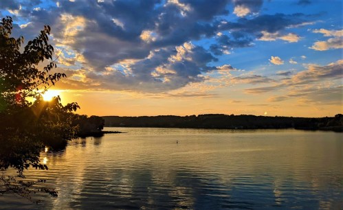 A serene lake at sunset, with vibrant clouds reflecting on the water and trees framing the scene.