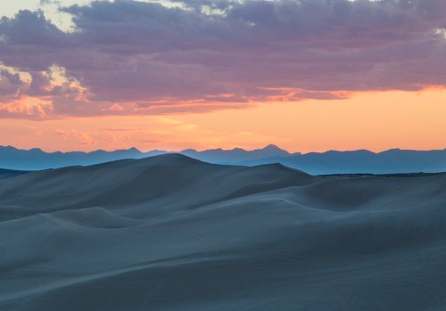 A serene desert landscape at sunset, with rolling sand dunes and distant mountains under a colorful sky.