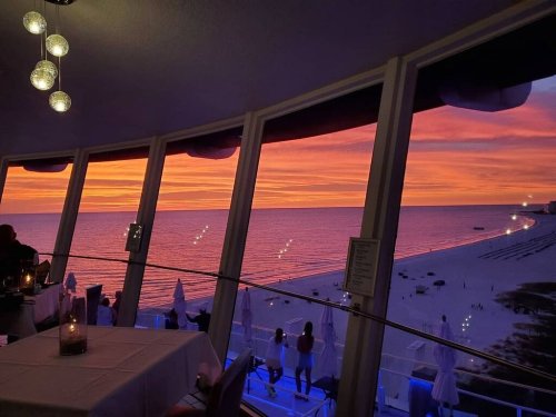 A stunning sunset over the ocean viewed from a restaurant, with silhouettes of people and beach umbrellas in the foreground.