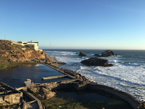 Coastal view featuring rocky formations, a sandy beach, and gentle waves under a clear blue sky.