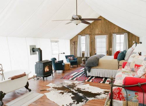 Cozy glamping tent interior with a bed, seating area, wood stove, and vintage decor, featuring a rustic charm.