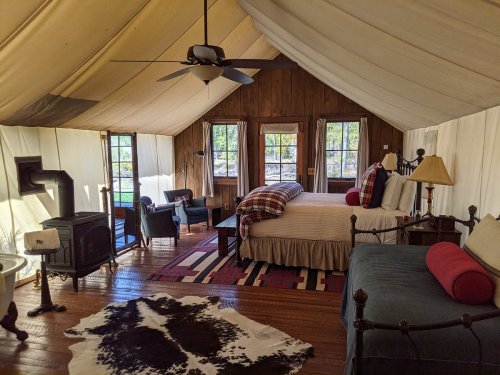 Cozy glamping tent interior with a bed, seating area, and rustic decor, featuring large windows and a wood stove.