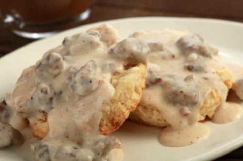 Two biscuits topped with creamy sausage gravy on a white plate. A glass of brown beverage is blurred in the background.
