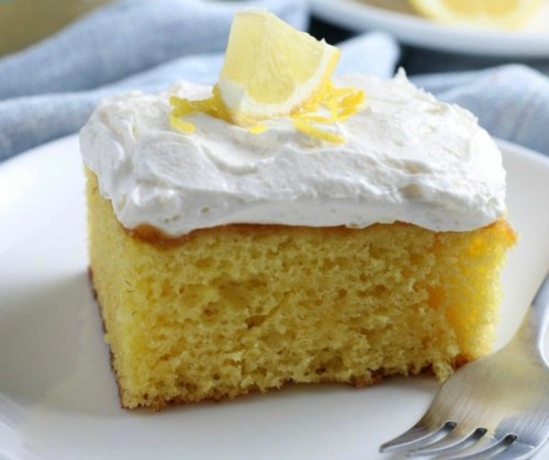 A slice of lemon cake topped with whipped cream and lemon zest on a white plate.