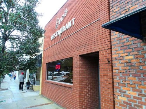 Exterior of a brick restaurant with a neon "OPEN" sign, located on a sidewalk with trees and pedestrians nearby.