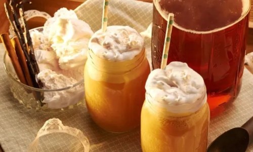 Two jars of creamy drinks topped with whipped cream, alongside a glass of iced tea and a bowl of dessert.