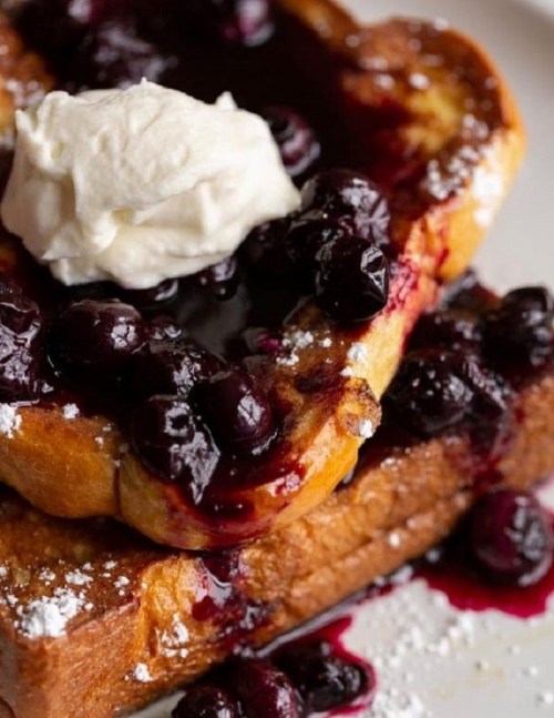 Close-up of French toast topped with blueberries, syrup, and a dollop of whipped cream on a white plate.