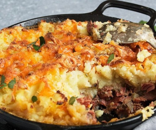 A cast iron skillet filled with cheesy mashed potatoes and meat, garnished with herbs, with a serving spoon beside it.