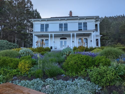 A large white house with a porch, surrounded by colorful gardens and trees in a serene landscape.