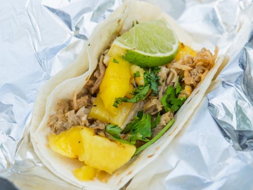 A taco filled with shredded meat, pineapple chunks, cilantro, and a lime wedge, wrapped in foil.