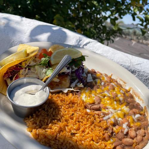 A plate of rice, beans with cheese, tacos, and a side of creamy sauce, set outdoors with greenery in the background.