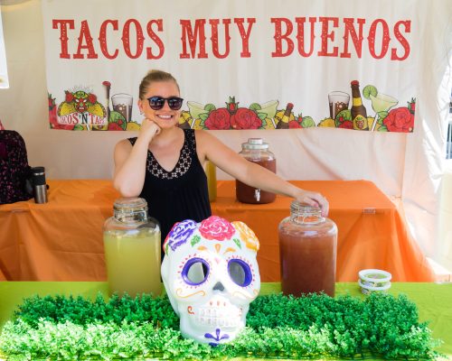 A smiling woman in sunglasses stands behind a table with drinks and a decorative skull, under a "Tacos Muy Buenos" banner.