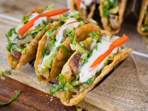 A close-up of crispy tacos filled with lettuce, meat, and topped with sauce and red pepper strips on a wooden surface.