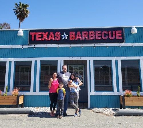A family poses in front of a blue building with a sign that reads "Texas Barbecue." Sunny day with palm trees.
