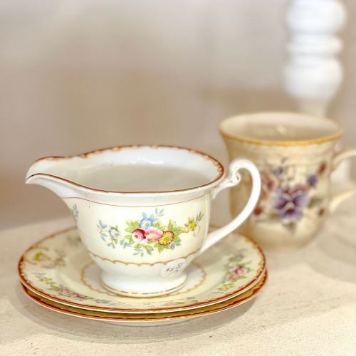 A delicate porcelain creamer with floral designs, placed on a matching plate, alongside an ornate cup.