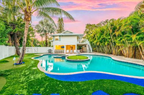 A beautiful backyard with a pool, palm trees, and a two-story house under a colorful sunset sky.
