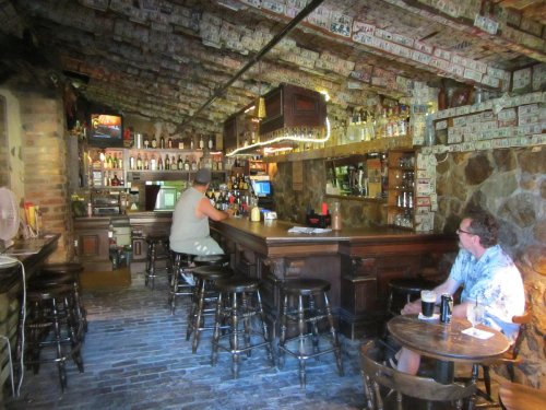 A rustic bar interior with a wooden counter, two patrons, and walls covered in dollar bills.