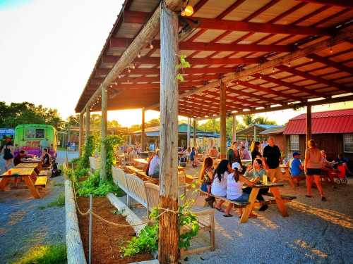 A lively outdoor market scene with people dining, food trucks, and a sunset backdrop under a wooden pavilion.