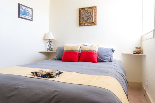 Cozy bedroom with a gray bedspread, red pillows, a lamp, and decorative wall art. Natural light fills the space.