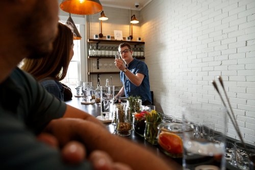 A bartender speaks to customers at a bar, surrounded by glassware and fresh ingredients on the counter.