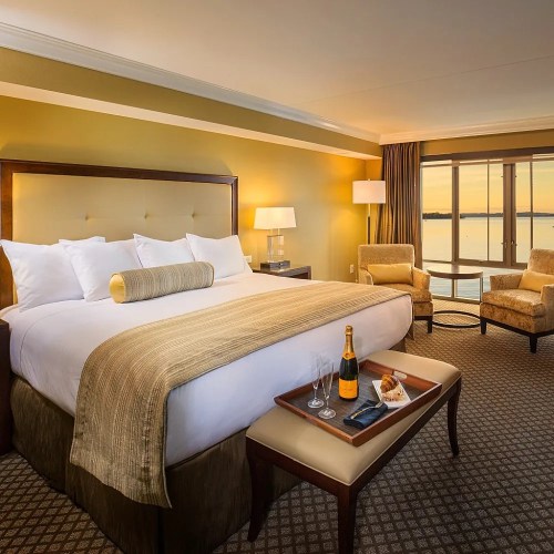 Luxurious hotel room with a king bed, seating area, and a view of the water at sunset. Champagne and snacks on a table.