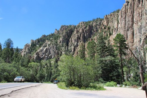 A scenic view of rocky cliffs and lush greenery alongside a road under a clear blue sky.