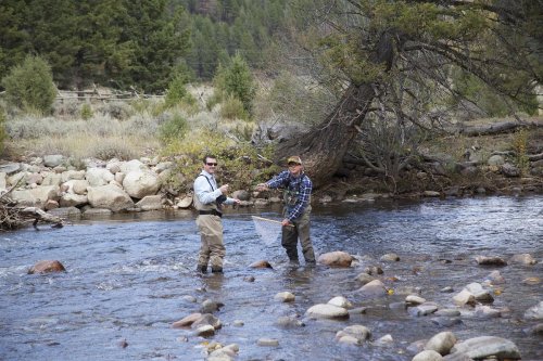 Two men fishing in a river, standing on rocks, with trees and a hillside in the background.