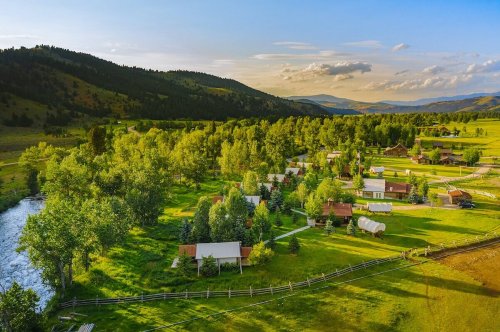 Aerial view of a serene countryside with cabins, trees, and a river, surrounded by rolling hills and mountains.