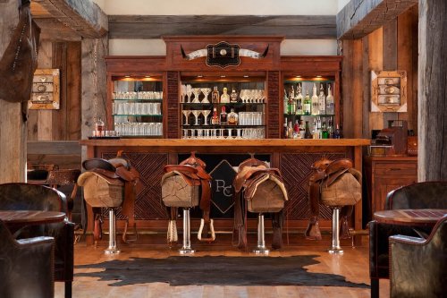 A rustic bar with saddle stools, wooden beams, and a variety of drinks displayed on shelves behind the counter.