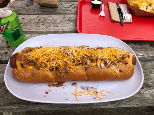 A large hot dog topped with cheese, onions, and chili, served on a white plate with a drink and fries in the background.