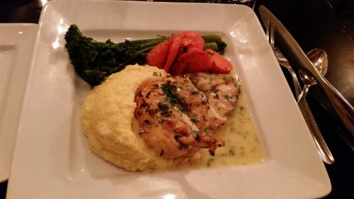 A plate featuring grilled chicken, creamy polenta, and sautéed broccoli and carrots, garnished with herbs.