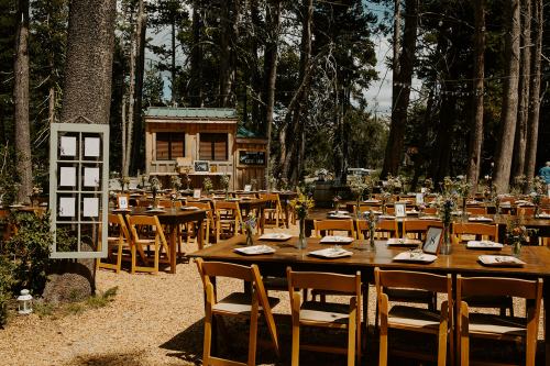 Outdoor dining setup with wooden tables and chairs, surrounded by trees, decorated with flowers and place settings.