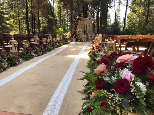 A beautifully decorated outdoor wedding aisle lined with flowers and lanterns, leading to a floral arch in a forest setting.