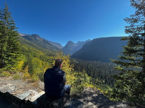 A person sits on a stone ledge, overlooking a vast mountain valley filled with trees under a clear blue sky.