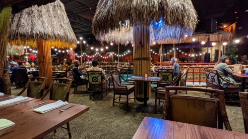 A tropical-themed restaurant interior with thatched roofs, dim lighting, and patrons seated at tables.