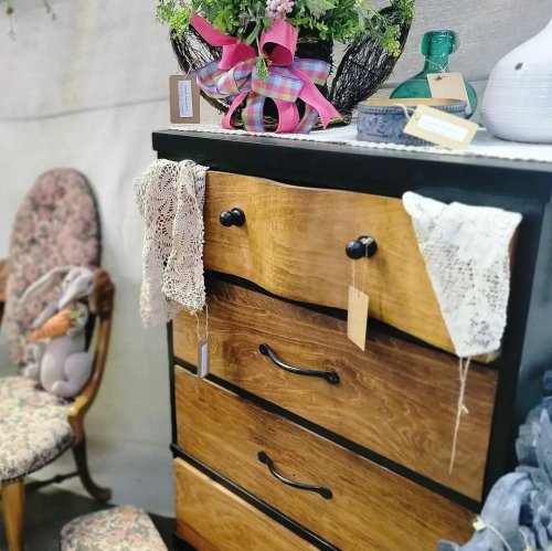 A wooden dresser with three drawers, decorated with lace and a floral arrangement, beside a vintage chair.